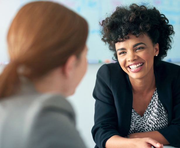 Two women are in a professional setting, possibly an office or a meeting room. One woman is in focus, smiling warmly and looking at the other woman, who is partially visible with her back to the camera. The woman in focus has a dark, curly hairstyle and is wearing a black blazer over a patterned top. The other woman, out of focus, has light-colored hair pulled back in a ponytail and is wearing a gray blazer. A whiteboard with some text and a colorful chart is visible in the blurry background.